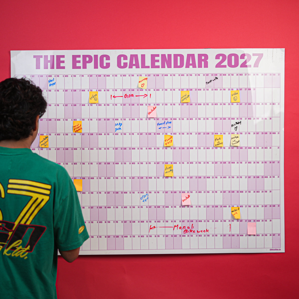 Person writing on a Rose EPIC Calendar mounted on a red wall